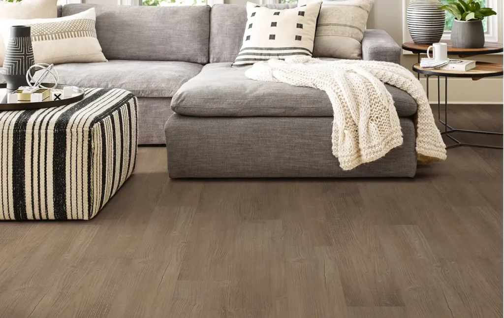 A cozy, contemporary living room featuring medium-toned, ash-brown wood-textured flooring. A large grey sectional sofa is styled with patterned throw pillows and a chunky cream knit blanket. To the side, a striped ottoman and a tiered black metal coffee table with potted plants add texture and a modern feel to the warm, inviting space.