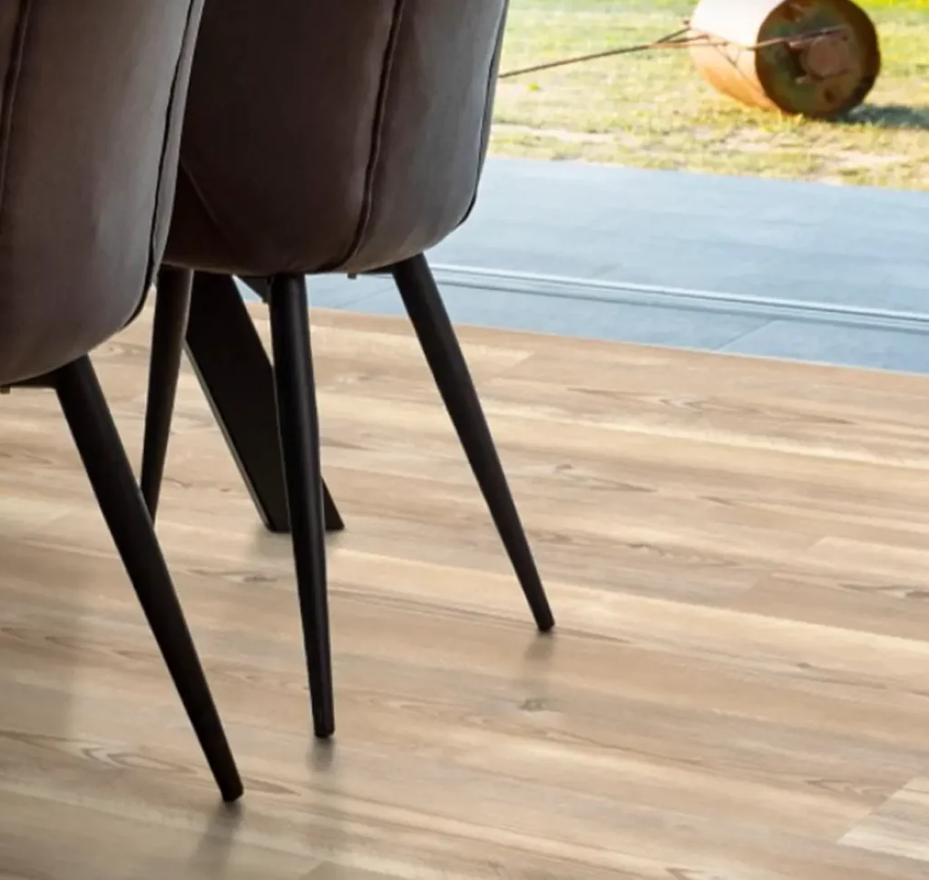 Close-up of light oak-style luxury vinyl plank flooring in a modern dining room with black-legged chairs and a view of an outdoor patio through a glass door.